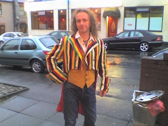 Andrew poses in a multi-coloured stripey tailcoat, hand on hip.