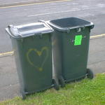 Two wheelie bins standing close together. One has a heart spray-painted on it.