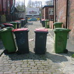 Four wheelie bins standing spread across a back alley, casting long shadows.