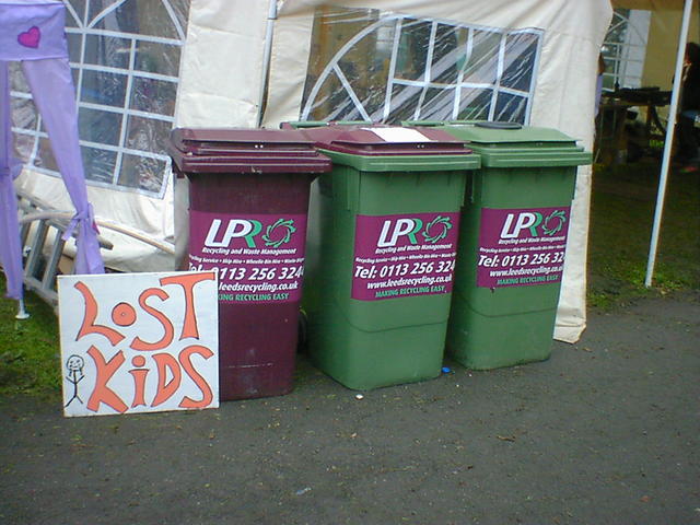 A sign for lost kids is leans next to 3 wheelie bins.
